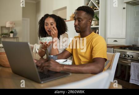 Ein junges, multiethnisches Paar, das zu Hause beim Frühstück an einem Laptop arbeitet Stockfoto