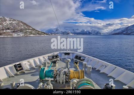 Blick über den Bug eines Hurtigruten Kreuzfahrtschiffs auf einem Fjord, umgeben von schneebedeckten Bergen, die sich Tromso, Norwegen nähern Stockfoto