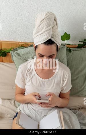 Eine junge Frau liegt im Bett mit einem weißen Handtuch um ihren Kopf gewickelt, während sie durch ihr Handy scrollt Stockfoto