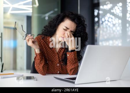 Übermüdete hispanische Frau bei der Arbeit, Geschäftsfrau mit Brille, die Hände am Kopf hält, hat starke Kopfschmerzen, arbeitet im Büro, sitzt am Tisch und benutzt ein Notebook bei der Arbeit. Stockfoto