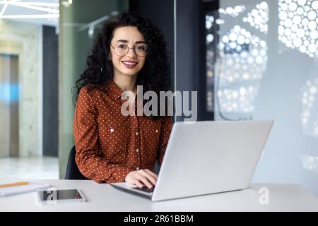 Junge, wunderschöne hispanische Frau, die in einem modernen Büro arbeitet, lächelt und mit einem Laptop in die Kamera schaut. Stockfoto