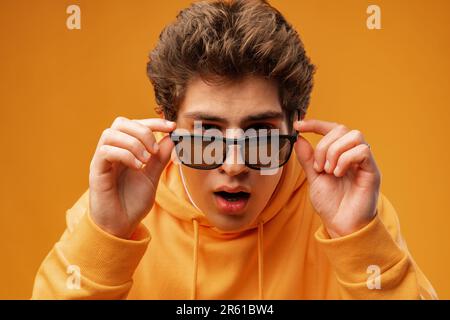 Attraktiver junger Mann posiert in Sweatshirt und Sonnenbrille auf gelbem Hintergrund Stockfoto