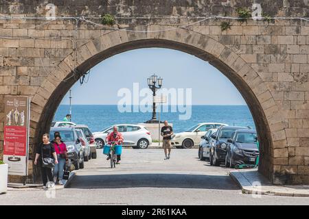 Touristen und Einheimische, die in der Nähe der alten Mauern mit einem großen Bogen, einer Straßenlampe und dem blauen Mittelmeer in der Altstadt von Bari, Apulien, Italien, spazieren gehen Stockfoto