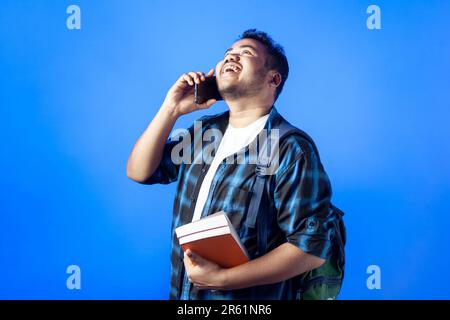 Porträt eines männlichen Studenten mit Büchern, der lacht und auf einem Smartphone spricht, mit Glück im blauen Hintergrund Stockfoto