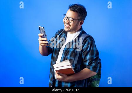 Glücklicher Mann, lächelndes Gesicht, indische ethnische Zugehörigkeit. Ein Studierender, der lächelt, während er sein Mobiltelefon mit Büchern benutzt Stockfoto