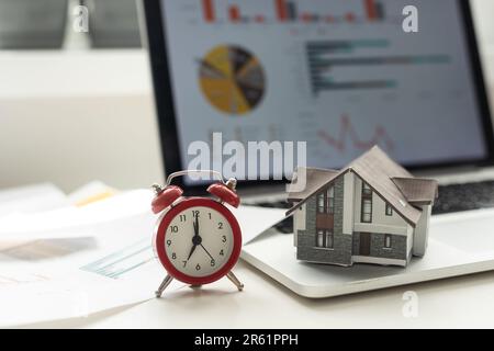 Wecker und Hausmodell. Zeit für eine Hypotheken- oder Immobilienversicherung Stockfoto