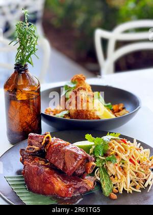 Eine saftige Platte mit gegrillten Schweinerippchen, begleitet von einem lebhaften grünen Papayasalat im thailändischen Stil, präsentiert auf einem weißen Teller Stockfoto