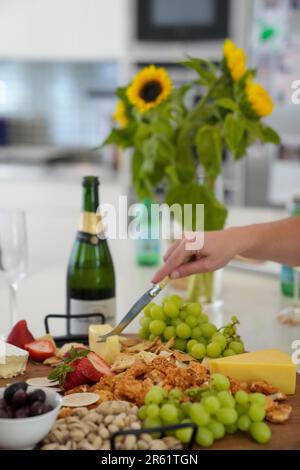Nahaufnahme einer Person, die einen Käseblock mit einem Messer schneidet, begleitet von Flaschen Wein und Tellern mit verschiedenen Speisen Stockfoto