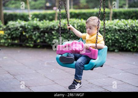 Der kleine blonde Junge mit Down-Syndrom reitet gern auf sicheren Kinderschaukeln Stockfoto