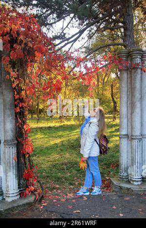 Auf dem Foto untersucht eine junge Frau in einer weißen Jacke die leuchtend roten Blätter wilder Trauben in einem Herbstpark. Stockfoto