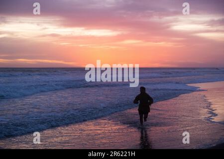 Eine Person, die in einer Silhouette vor einem lebhaften Hintergrund läuft, in dem die untergehende Sonne von der Oberfläche des Ozeans reflektiert wird Stockfoto