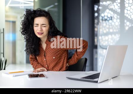 Wunderschöne lateinamerikanische Geschäftsfrau, die in modernen Büros arbeitet, übermüdete Arbeiterin, hat starke Rückenschmerzen, wenn sie ein Notebook benutzt. Stockfoto