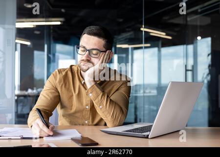 Gelangweilter und trauriger junger Mann, der im Büro an einem Laptop arbeitet und sich mit einem Stift Notizen in einem Notizbuch macht. Überarbeitet, müde von der Arbeit. Stockfoto