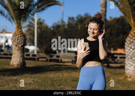 Lächelnde junge Frau macht beim Training Pause, benutzt Smartphone, hört Musik über Kopfhörer, trägt Sportkleidung im Freien auf Palmen-Hintergrund. Welt-er Stockfoto