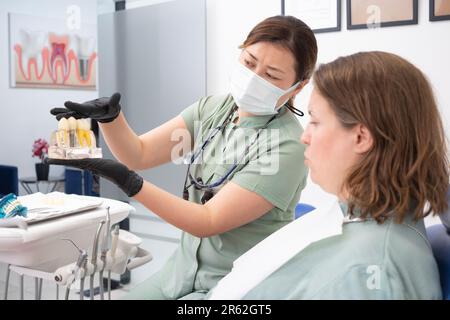 Zahnarzt, der einem Patienten ein Modell menschlicher Zähne hält, erklärt den Prozess der Implantatplatzierung, Stockfoto