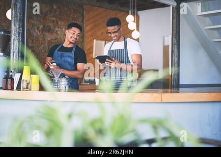 Wer braucht Papier, um ein profitables Geschäft zu führen? Zwei junge Männer, die ein digitales Tablet verwenden, während sie Kaffee in einem Café zubereiten. Stockfoto