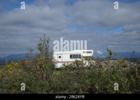 Ein Wohnmobil, das inmitten von weiten Landschaften, grünen Ebenen und ruhigen und entfernten Bergen parkt und den abgelegenen ruhigen Ort unter blauem und bewölktem Himmel erreicht. Stockfoto