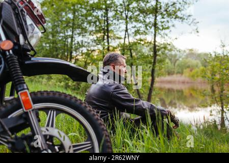 Porträt eines erfahrenen Motorradfahrers, der sich im Freien mit dem Motorrad entspannt. Ein Mann mit Lederjacke genießt die Flusslandschaft. Ältere Motorradfahrer, die surren Stockfoto