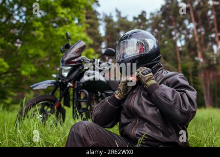 Nahaufnahme des Motorradfahrers, der den Helm nach dem Motorradfahren im Sommerwald abnimmt. Ein Mann, der draußen Lederkleidung trägt. Sicherheitsmaßnahmen Stockfoto
