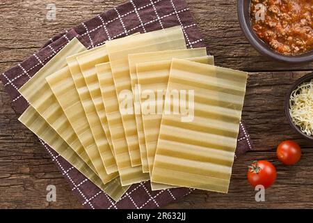 Trockene, ungekochte, gerippte Lasagne-Pasta-Blätter, fotografiert über dem Kopf auf rustikalem Holz mit Bolognese-Sauce, geriebenem Käse und Kirschtomaten an der Seite Stockfoto