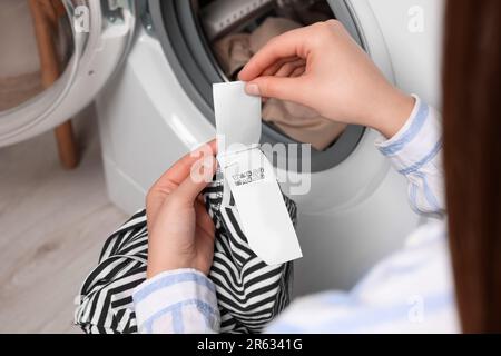 Frau liest Kleidungsetikett mit Pflegesymbolen und Materialinhalt auf gestreiftem Hemd in der Nähe der Waschmaschine, Nahaufnahme Stockfoto