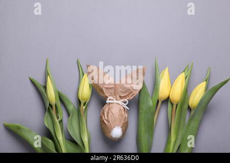 Osterhasen aus Kraftpapier und Ei neben schönen Tulpen auf grauem Hintergrund, flach liegend. Platz für Text Stockfoto