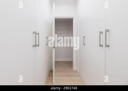 Ein begehbarer Schrank mit weißen Holztürenschränken auf beiden Seiten und einem Holzboden mit einer offenen weißen Tür und grauen Metallzügen Stockfoto