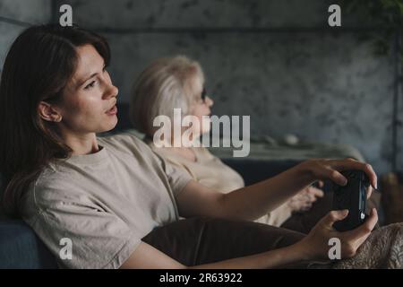 Fröhliche zwei weiße Frauen, die zu Hause zusammen bleiben, Gamepads benutzen, auf den Bildschirm schauen und lächeln. Unterhaltung mit digitaler Technologie. Familie Stockfoto