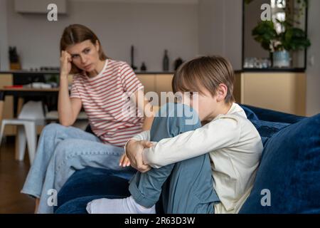 Ein frustrierter Teenager, der die Knie auf der Couch umarmt und wegschaut. Mama sitzt neben dem Trost-Sohn Stockfoto