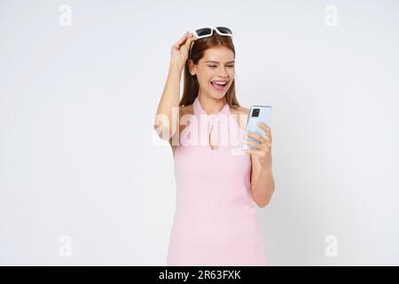 Junge Frau, die ein Smartphone in der Hand hält und lächelt und eine Sonnenbrille trägt, isoliert auf weißem Hintergrund. Stockfoto