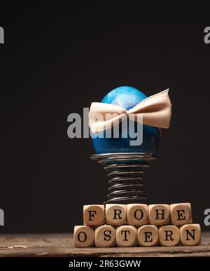 Osterei mit einem Bogen auf einem alten rustikalen Holztisch, Holz- Würfel mit der Deutschen Wörter Frohe Ostern Stockfoto