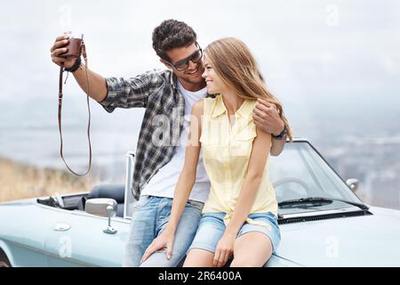 Sagen Sie Cheese. Ein glückliches junges Paar, das während eines Roadtrips in seinem Cabrio Fotos macht. Stockfoto