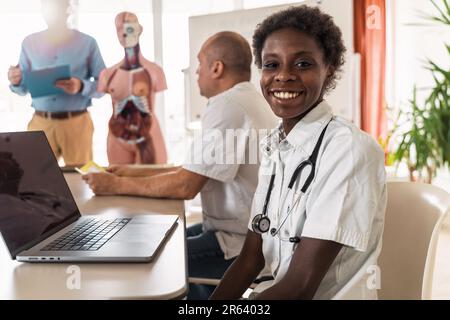 Afrikanische Ärztin leitet engagierten Workshop in Kliniktagungsraum. Sie lächelt, während sie mit Kollegen interagiert, mit einem Laptop in der Hand. Vielfältiger medizinischer Profi Stockfoto