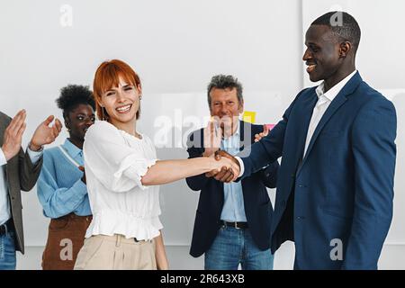 Ein Moment des Erfolgs und der Freude, während eine junge rothaarige kaukasische Frau einem afrikanischen Mann in einem schwarzen Anzug die Hand schüttelt, während ihre vielfältigen Kollegen zuschlagen Stockfoto