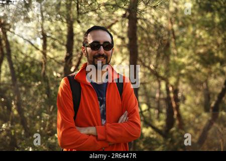 Ein bärtiger Mann mit Sonnenbrille und ein orangefarbenes Hemd, der vor einem unscharfen grünen Hintergrund im Wald posiert Stockfoto