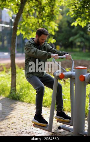 Bärtiger weißer Mann mit grauer Sportkleidung, der öffentliche Trainingsgeräte im Park benutzt Stockfoto