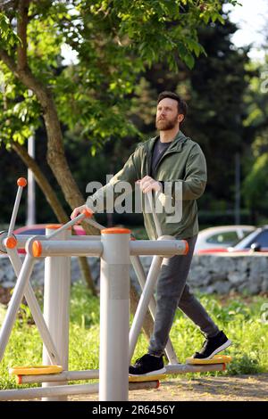 Bärtiger weißer Mann mit grauer Sportkleidung, der öffentliche Trainingsgeräte im Park benutzt Stockfoto