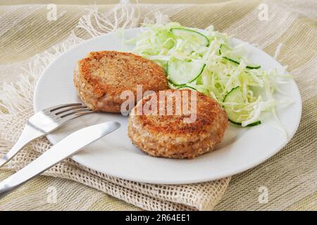 Fischkuchen oder Koteletts. Aus Erdbarsch und Thunfisch mit Kräutern, paniert und gebraten, serviert mit Salat auf einem weißen Teller Stockfoto