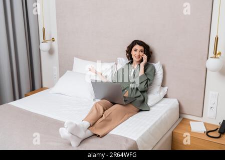 Die ganze Länge voller fröhlicher tätowierter Frau in lässiger Kleidung, die auf dem Bett sitzt, mit einem Laptop neben weißen Kissen, Wandleuchtern und grauen Vorhängen, Gesten während der Fahrt Stockfoto