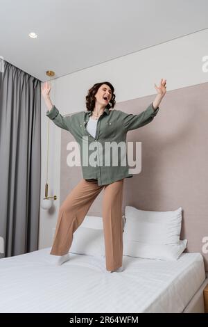 Lange junge und überfreuliche Frau in lässiger Kleidung und mit welligen braunen Haaren, die tanzen und singen auf dem Bett neben weißen Kissen, grauen Vorhängen und Stockfoto