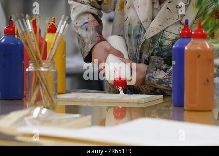 Ein Künstler oder Student visueller Kunst im Areal spritzt oder gießt Acrylfarbe aus einer weißen Flasche auf eine Malpalette. Organisiertes Klassenzimmer. Ar Stockfoto