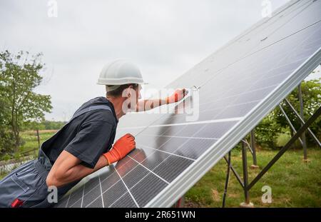 Mann Ingenieur Installation von Solarmodulen. Männlicher Arbeiter in Schutzhelm und Arbeitshandschuhen, die die mittlere Klemme während der Montage des Solar-Photovoltaik-Panelsystems überprüfen. Stockfoto