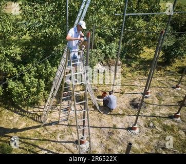 Arbeiter steht auf Treppe und schneidet Metallbalken mit Schleifer zur Installation von Solarpaneelen. Bodenmontierte Photovoltaiksysteme. Solarmodule werden durch Racks oder Rahmen an Ort und Stelle gehalten. Stockfoto