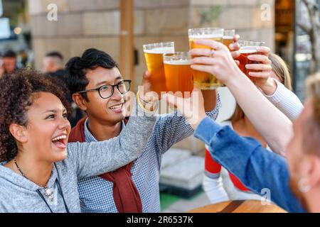 Gruppe positiver multiethnischer Freunde mit männlichem Crop in legerer Kleidung, lächelnd und schauend zu Biergläsern und klimpernd, während er auf hölzernem Ta steht Stockfoto