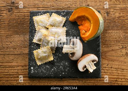 Blick von oben auf ungekochte italienische Ravioli auf Schieferbrett mit aromatischen Pilzen und frischem Kürbis auf einem Holztisch Stockfoto