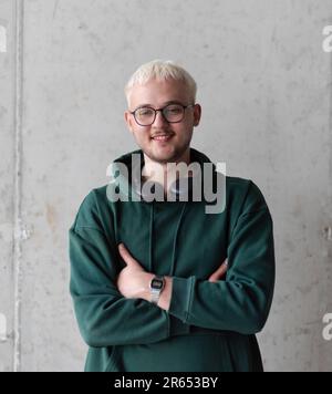 Ein Mann mit blauen Haaren, einer Brille und einem grünen Sweatshirt posiert selbstbewusst mit gekreuzten Armen vor grauem Hintergrund und zeigt sein modisches Gesicht Stockfoto