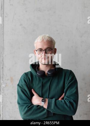 Ein Mann mit blauen Haaren, einer Brille und einem grünen Sweatshirt posiert selbstbewusst mit gekreuzten Armen vor grauem Hintergrund und zeigt sein modisches Gesicht Stockfoto