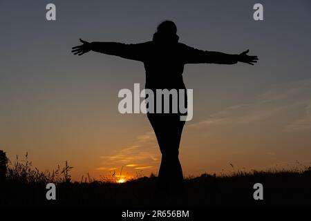 Die Silhouette der Rückansicht einer Frau, die über ein Feld in Richtung Sonnenuntergang in Großbritannien blickt, mit ausgestreckten Armen. Stockfoto
