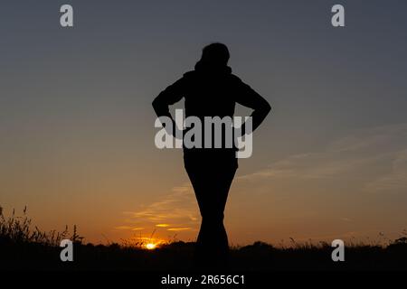 Rückansicht einer Frau, die über ein Feld in Richtung Sonnenuntergang blickt, ihre Hände auf der Hüfte. Stockfoto