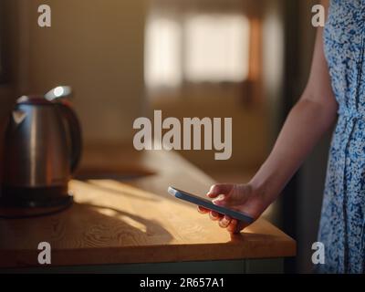 Weibliche Hand mit Smartphone in der Küche, mit Morgensonne. Winzige Häuser und kleines Wohnkonzept. Stockfoto